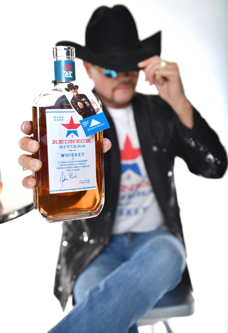 John Rich in sunglasses and a cowboy hat holding out a bottle of Redneck Riviera Whiskey