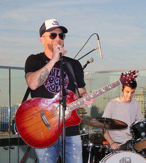 A LOCASH singer in sunglasses and a cap sings with a red acoustic guitar on a rooftop stage