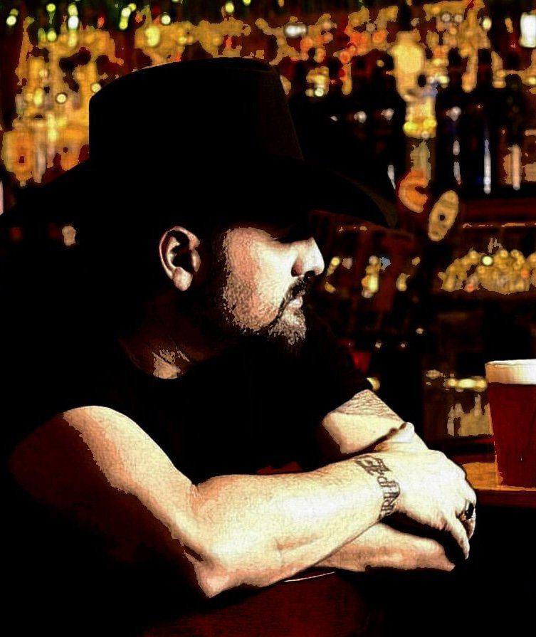 Ray Scott in a black hat leaning on a bar, a glass of beer beside him