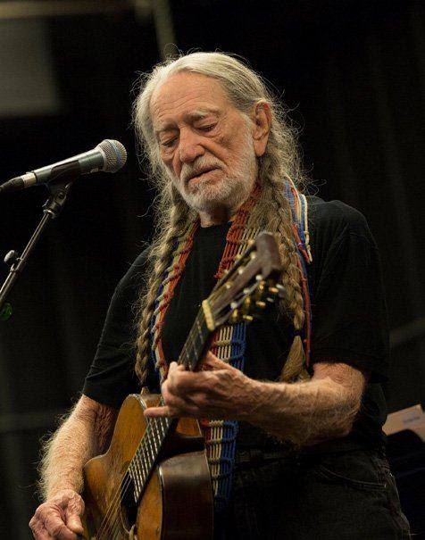 Willie Nelson playing guitar and singing into a microphone on stage