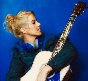 Alyssa Jacey with blonde hair and blue headphones holding an acoustic guitar against a blue background