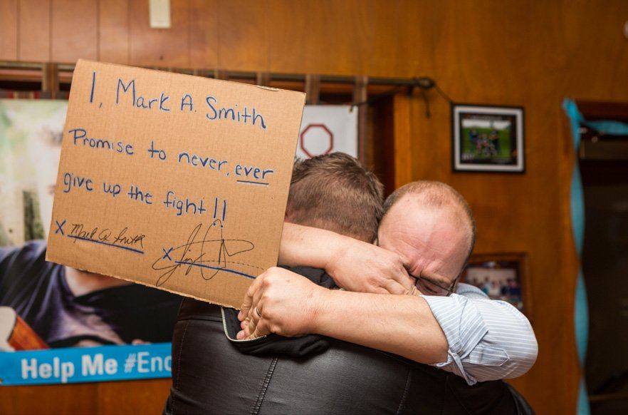 Two men embracing beside a handwritten sign reading “I, Mark A. Smith Promise to never ever give up the fight!!”
