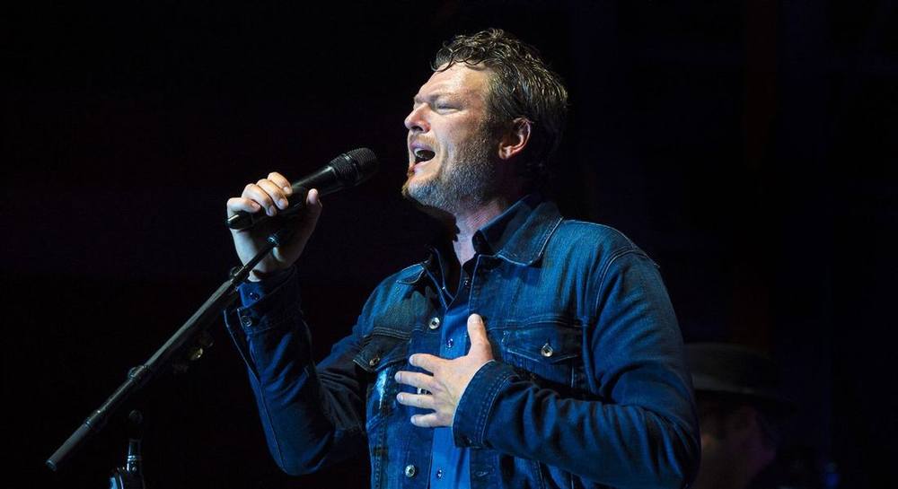 Blake Shelton singing into a microphone on stage in a denim jacket