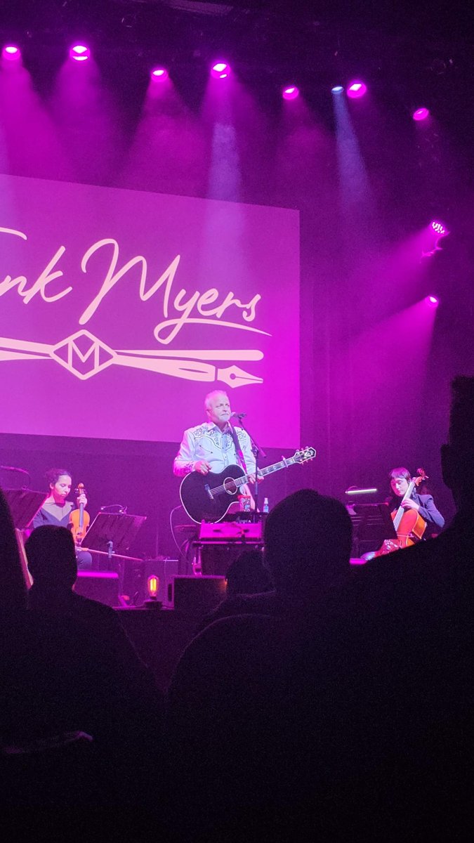 Frank Myers on stage with string players under purple lights, his name on the screen behind him